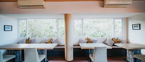 Un comedor con mesas y bancos de madera, ventanas amplias con vistas a vegetación y unidades de aire acondicionado en el techo.
