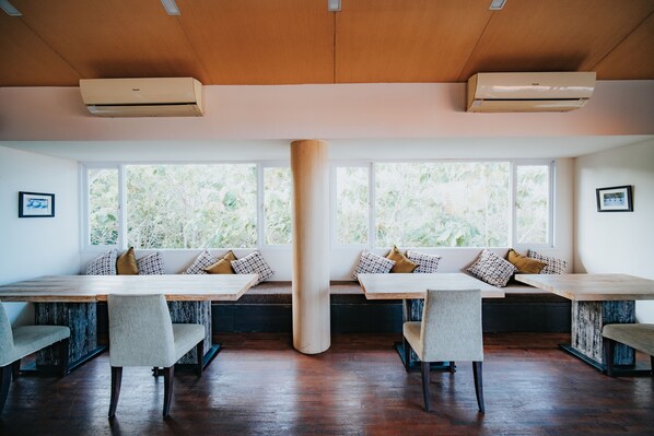 Un comedor con mesas y bancos de madera, ventanas amplias con vistas a vegetación y unidades de aire acondicionado en el techo.