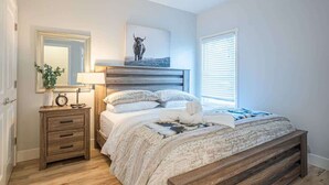 Un dormitorio con estructura de cama de madera, una mesita de noche con lámpara, un espejo y un cuadro enmarcado en la pared.
