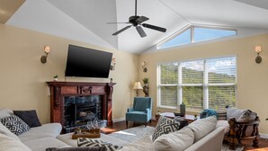 Una sala de estar con chimenea, ventilador de techo, televisor grande y vistas al exterior a través de las ventanas.
