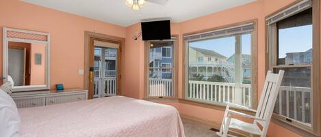 A bedroom with a bed, a rocking chair, a ceiling fan, and large windows with a view of a balcony and buildings.