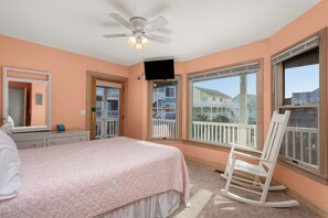 Une chambre à coucher comprenant un lit, un fauteuil à bascule, un ventilateur de plafond et de grandes fenêtres donnant sur un balcon et des immeubles.