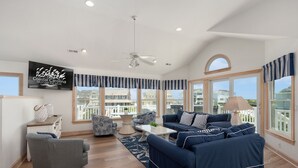 A spacious living room with a large sofa, armchairs, and a coffee table. A television is mounted on the wall, and there are large windows offering a view of the surrounding area. - C-Side: Semi-oceanfront, hot tub, community oceanfront pool, walkway to beach by house. (Nags Head)