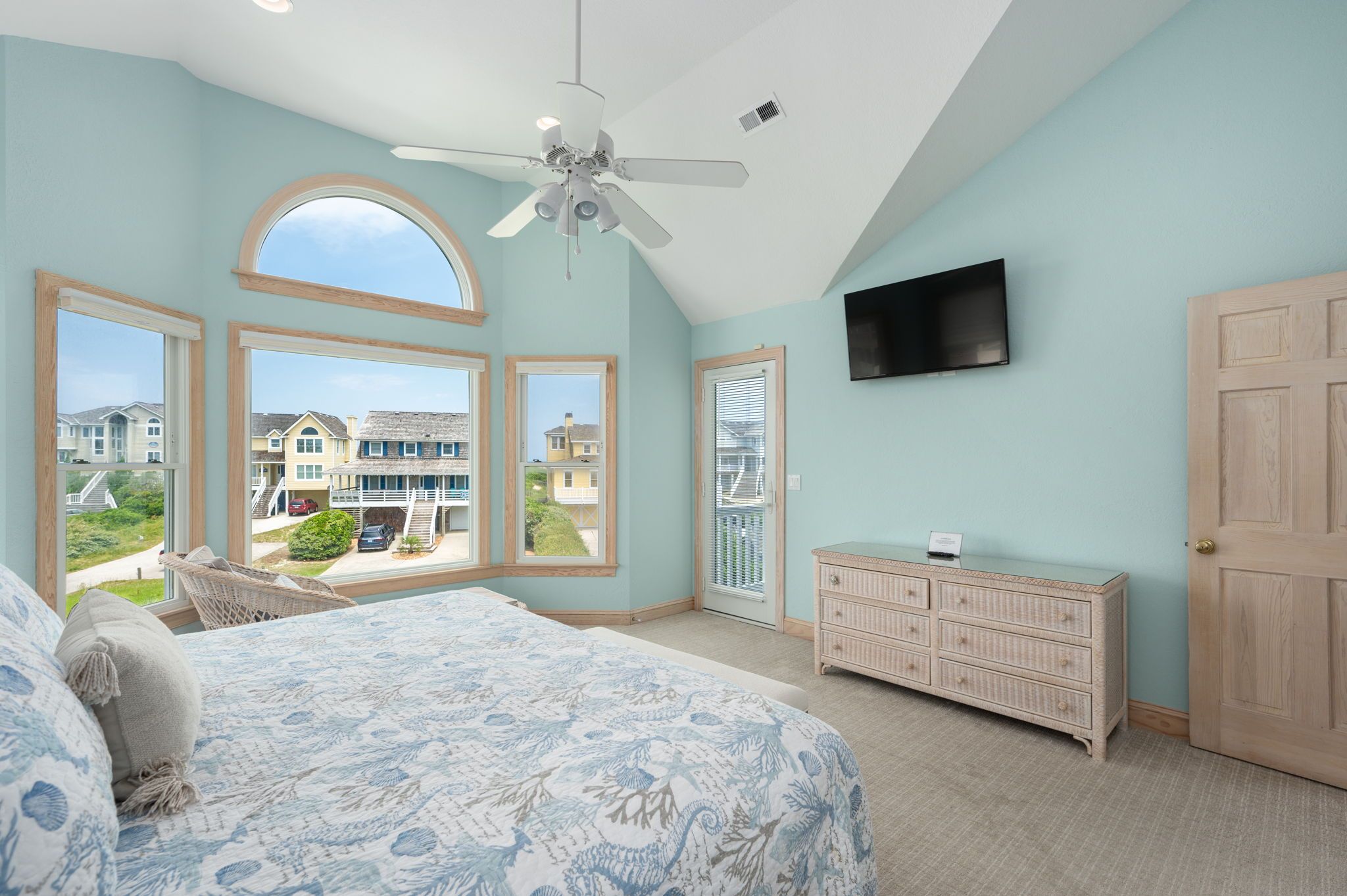 A bedroom with a large bed, a ceiling fan, a TV, and a view of houses through large windows.