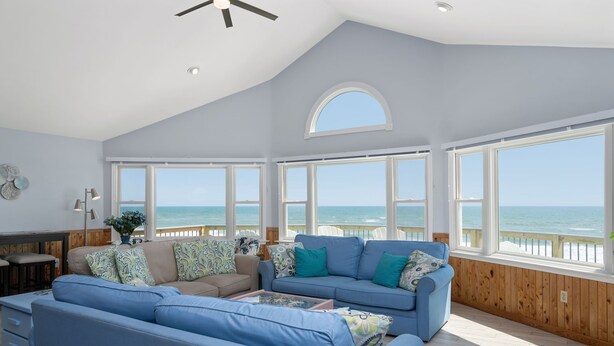 A spacious living room with a large window overlooking the ocean, a ceiling fan, and a wooden floor. - SeaAira: Oceanfront with unbeatable ocean views, renovated kitchen and baths (Nags Head)