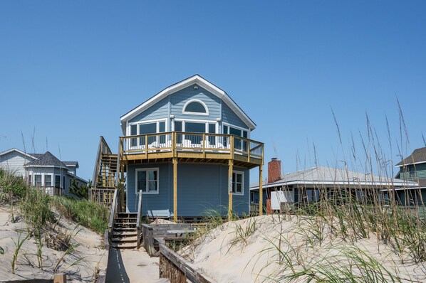A two-story beach house with a balcony, a staircase leading to the entrance, and a sandy beach in the background.