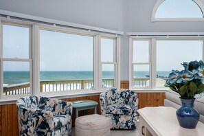 A room with a view of the sea, featuring a wooden bench, two chairs, a small table, and a vase with flowers.