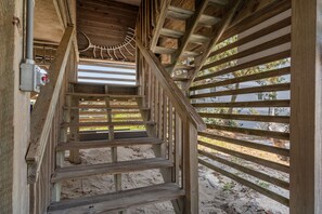 A wooden staircase leading to a covered outdoor area with a view of a sandy beach and a building in the background.