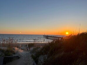 A beach scene with a wooden pier extending into the water, a sandy path leading to the water, and a clear sky.