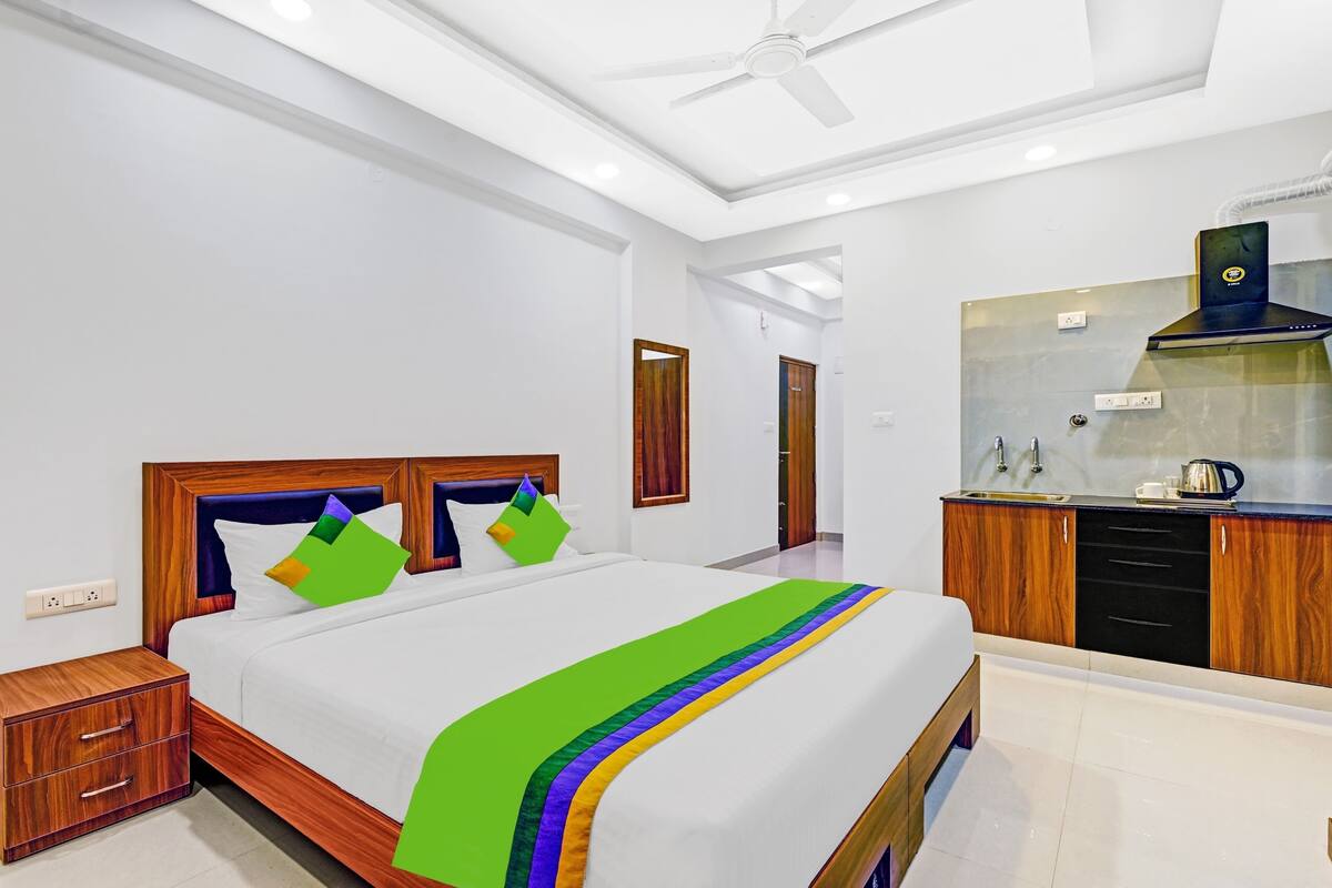 Una habitación de hotel moderna con una cama grande, una mesita de noche de madera, un ventilador de techo, una kitchenette y un baño visible a través de una puerta abierta.