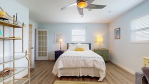 A bedroom with a bed, a ceiling fan, a window with blinds, a bedside table with a lamp, and a shelf with decorative items. - The Regal Beagle: 2  BR, 2  BA House in Crystal Beach, Sleeps 8 (Bolivar Peninsula)