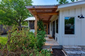 Eine überdachte Terrasse mit Steinstufen, die zu einem weißen Gebäude mit großen Fenstern und einer Pergola darüber führen.
