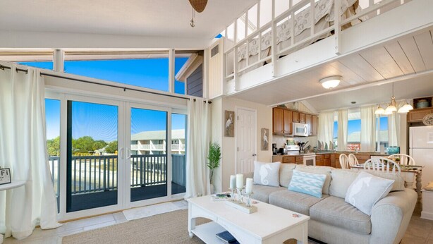 A spacious living area with a large sliding glass door leading to a balcony, a white sofa set, a kitchen with wooden cabinets, and a dining area with white chairs.