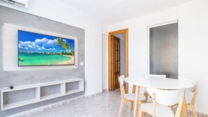 Une chambre moderne avec un grand téléviseur fixé au mur, diffusant une scène de plage, une table ronde blanche avec des chaises et une porte en bois donnant sur une autre pièce.