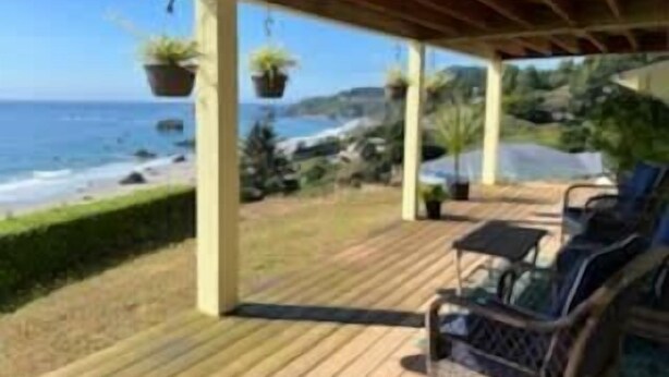 Terraza de madera con mobiliario exterior y macetas colgantes con vistas a una playa.