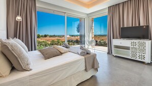A modern bedroom with a large bed, a TV on a white cabinet, and a view of the landscape through large windows.