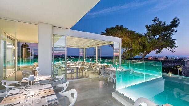 An outdoor dining area with tables and chairs, a pool, and a view of the city at dusk.