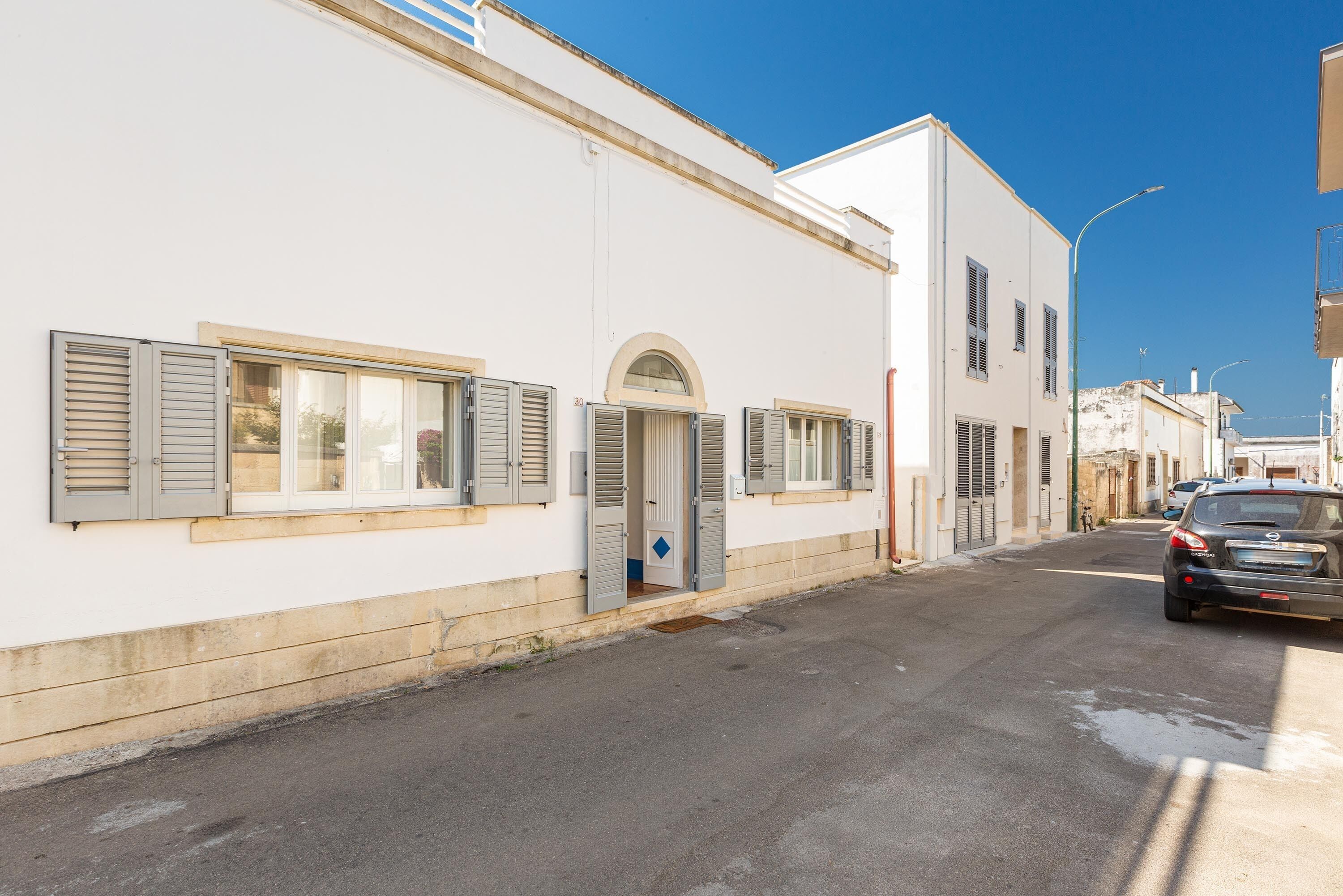 Un carrer estret amb edificis blancs, persianes a les finestres i un cotxe estacionat.