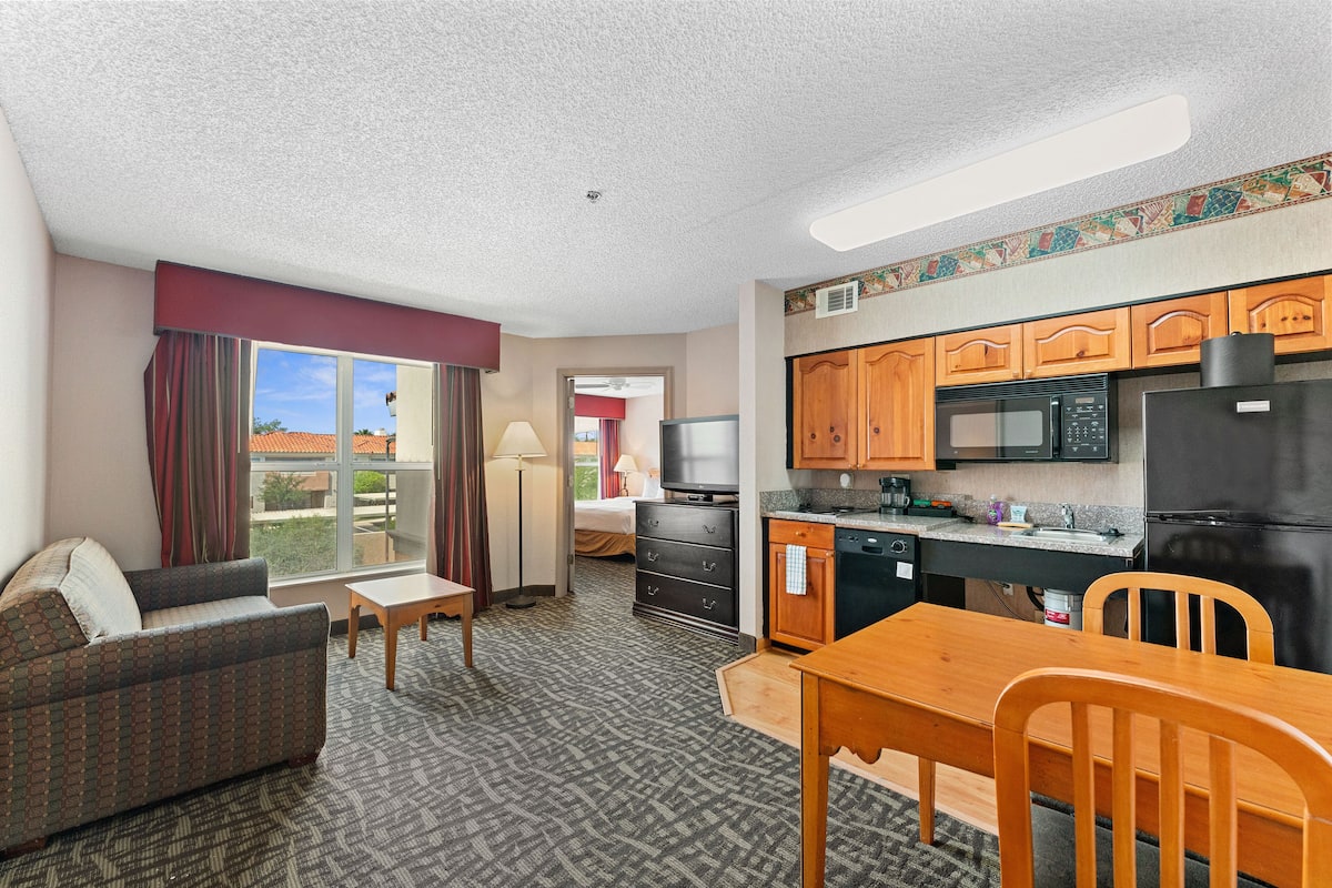 A compact hotel room with a kitchenette, a sofa, a dining table, and a TV.