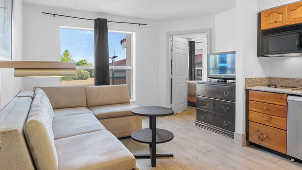 A modern living room with a beige sectional sofa, a small round table, and a kitchen area with wooden cabinets and a built-in microwave. - Metro Scottsdale Extended Stay (Paradise Valley)
