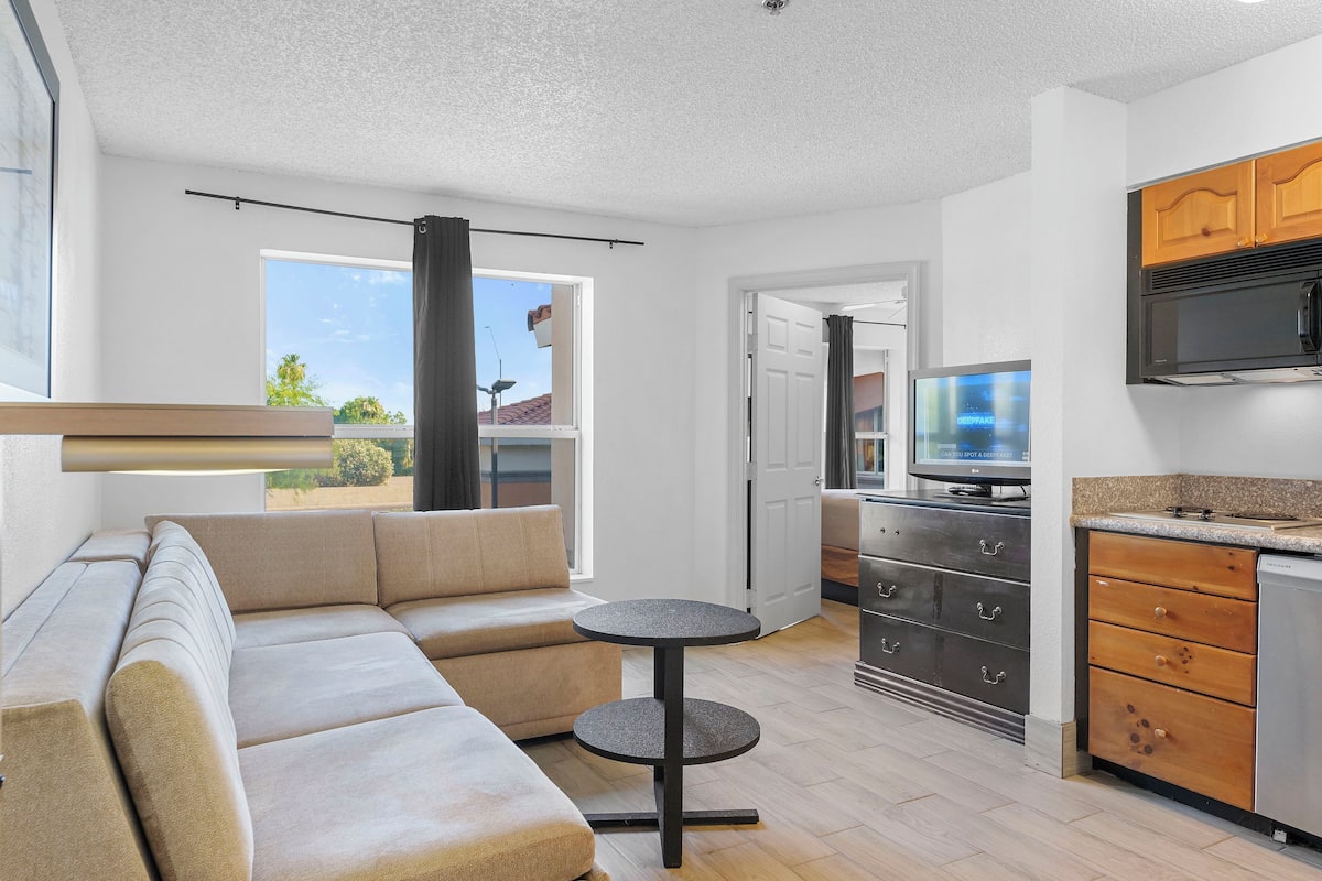 A modern living room with a beige sectional sofa, a small round table, and a kitchen area with wooden cabinets and a built-in microwave.