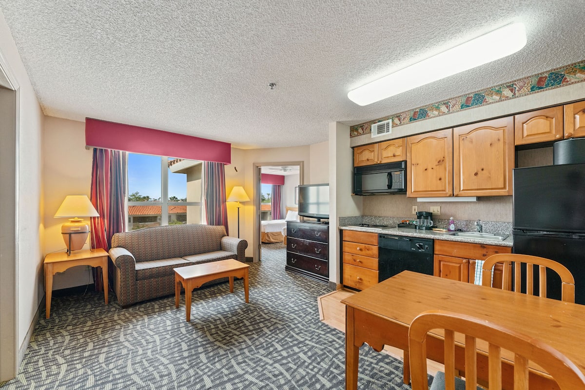 A compact hotel room with a kitchenette, a sofa, a dining table, and a TV.