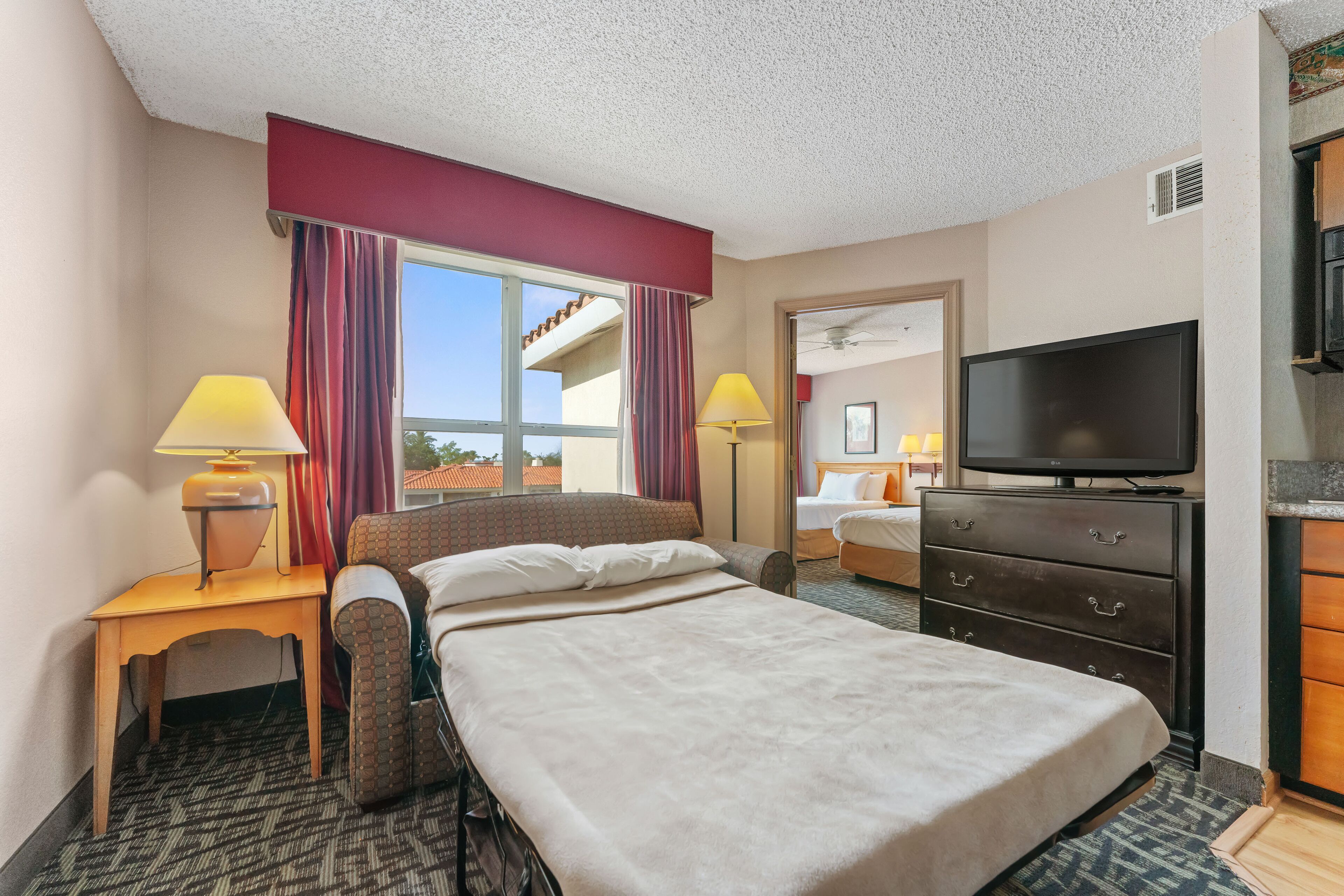 A hotel room with a bed, a desk with a lamp, a television, and a view of the outside.
