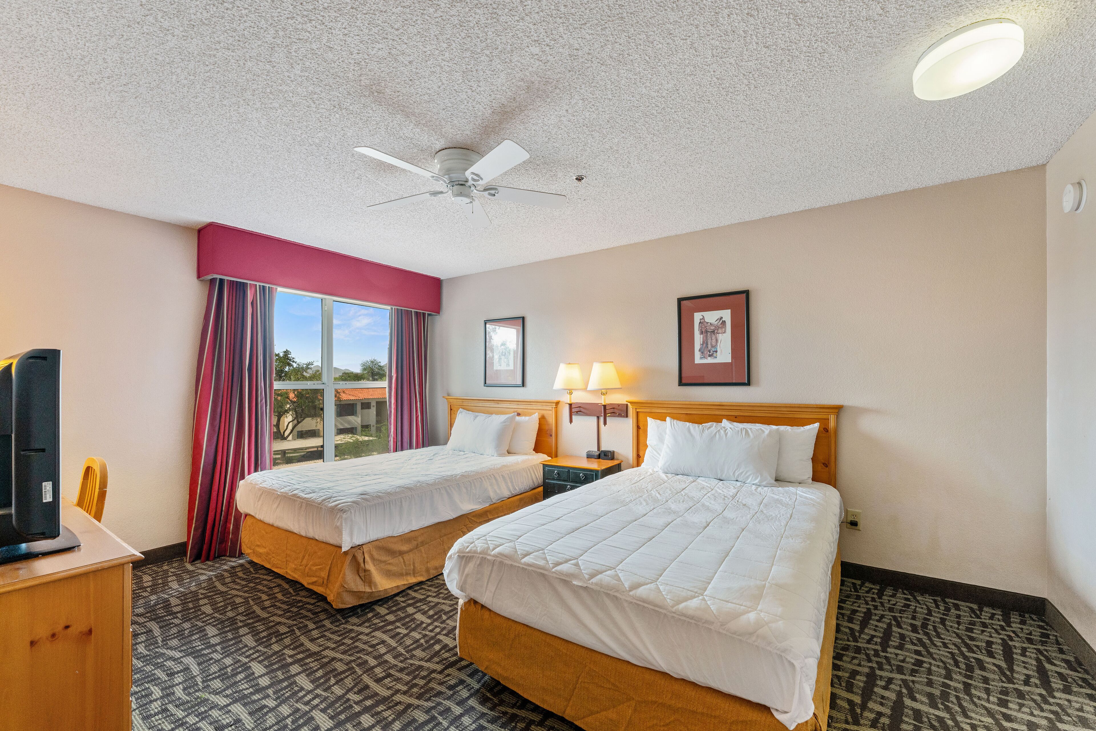 A hotel room with two beds, a ceiling fan, a TV, and a window with curtains.