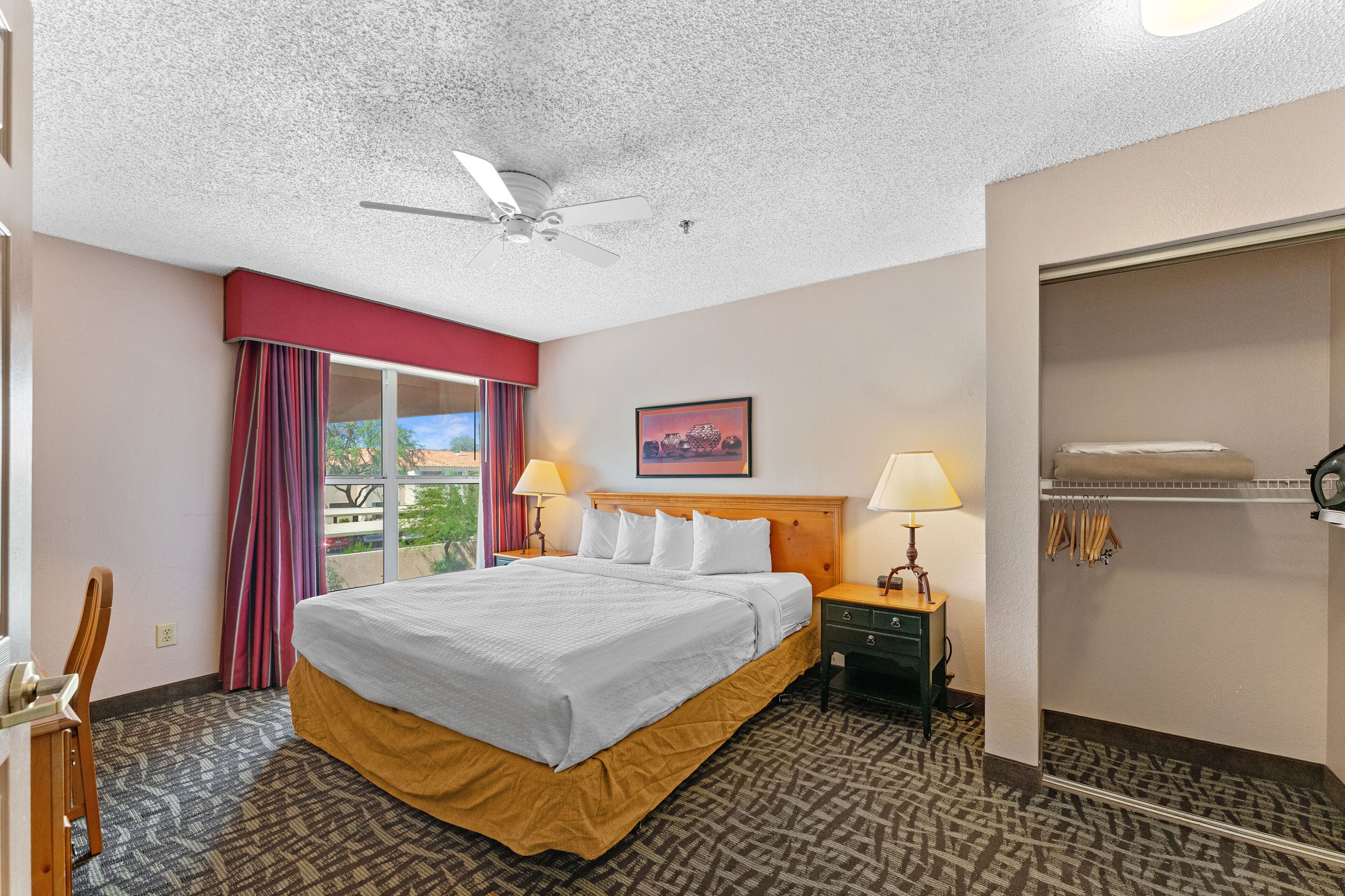 A hotel room with a bed, a nightstand, a lamp, a ceiling fan, a window with curtains, and a view of the outdoors.