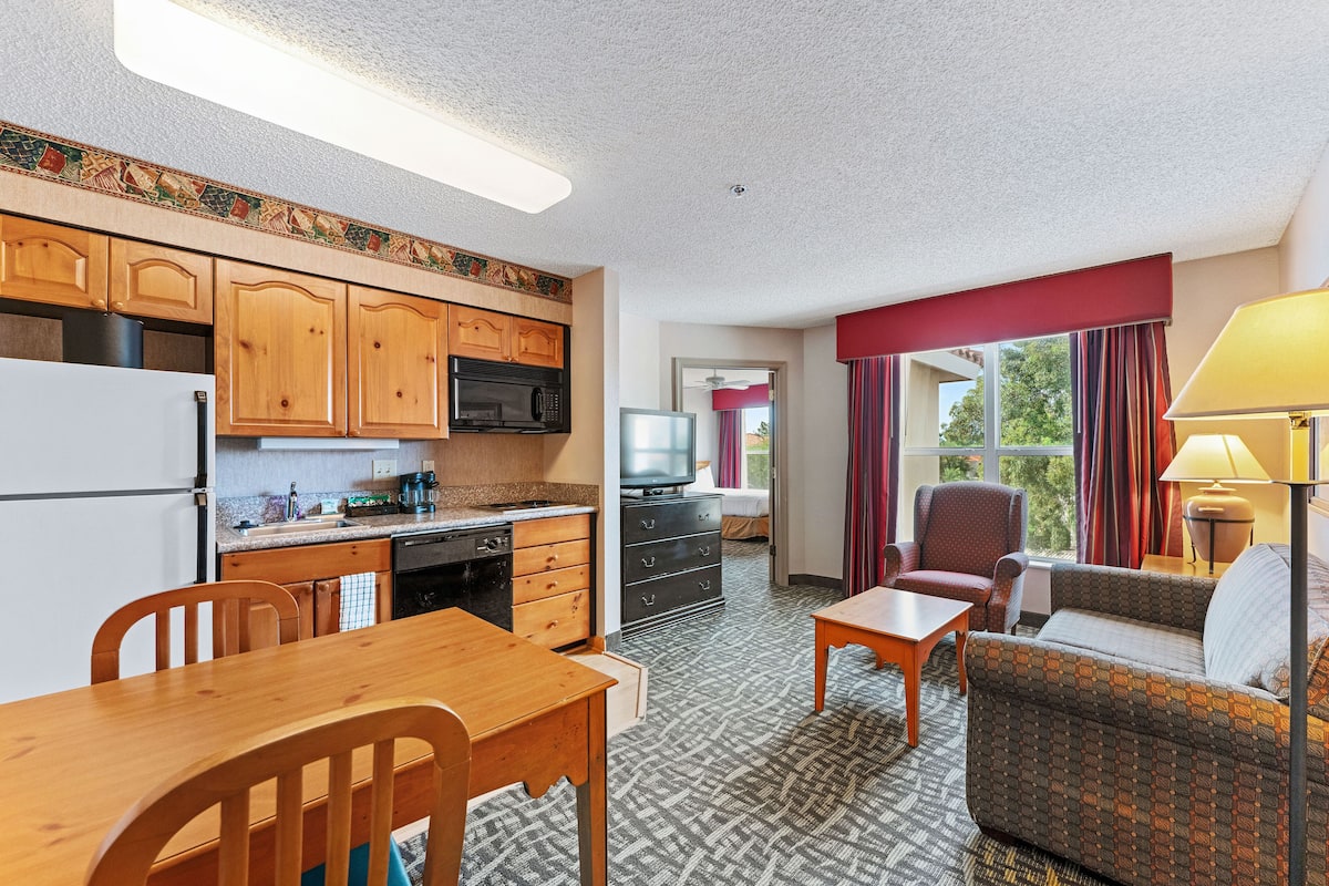A compact hotel room with a kitchenette, a dining area, a sofa, and a TV.