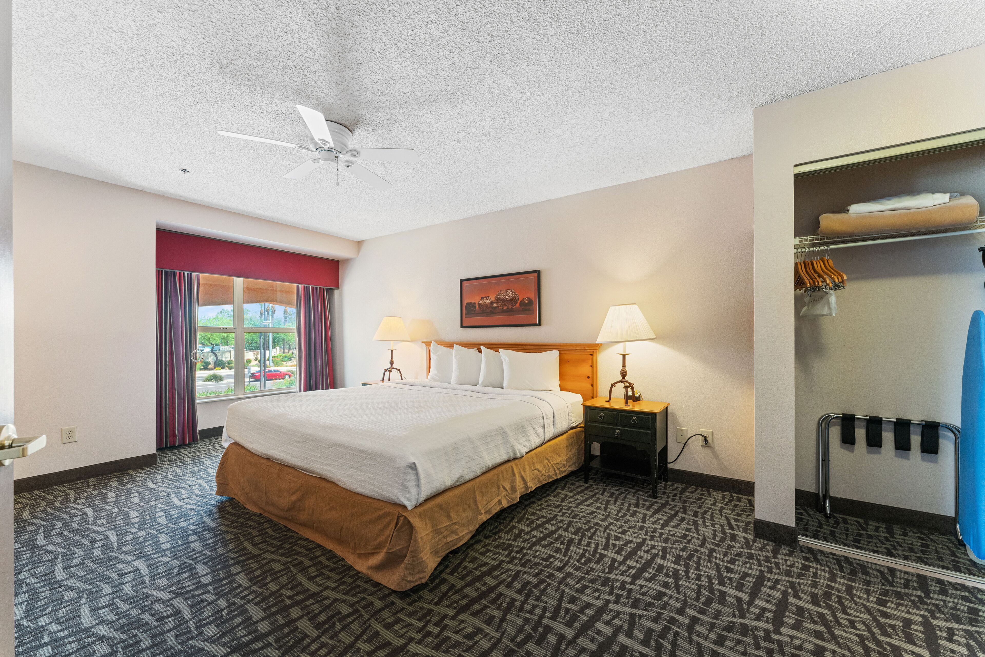 A hotel room with a bed, two bedside lamps, a ceiling fan, a window with curtains, a wall-mounted coat rack, and a door with a mirror.