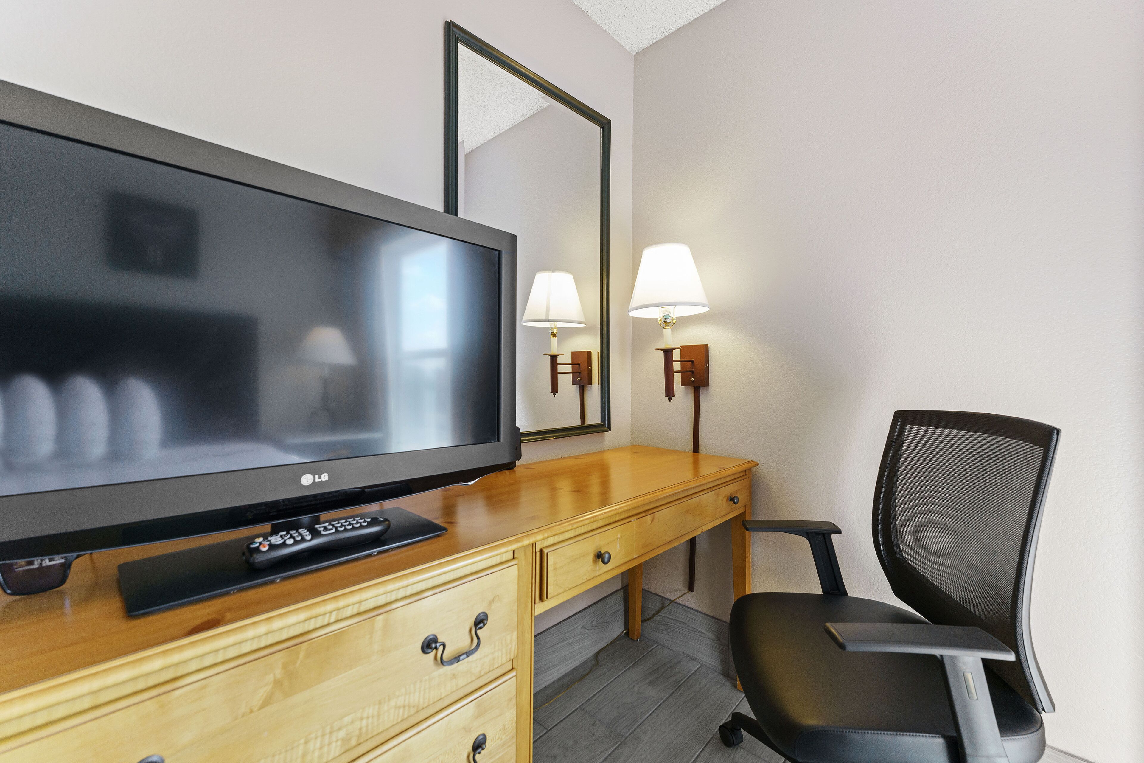 Habitación de hotel con televisor de pantalla plana, escritorio de madera, dos cajones, una silla y dos lámparas fijadas a la pared.