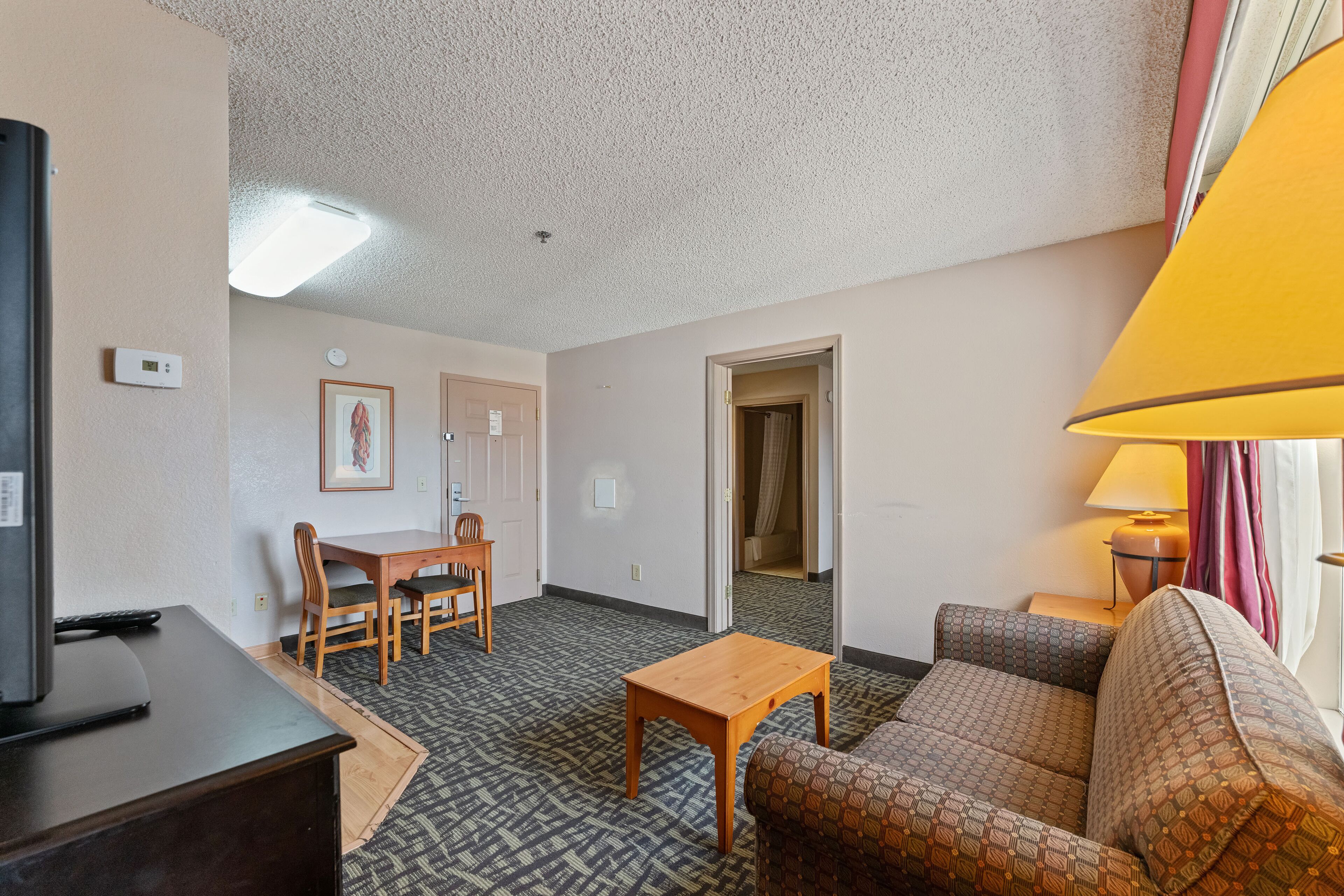 A hotel room with a sofa, a small wooden table, a TV on a stand, a dining table with chairs, and a lamp.