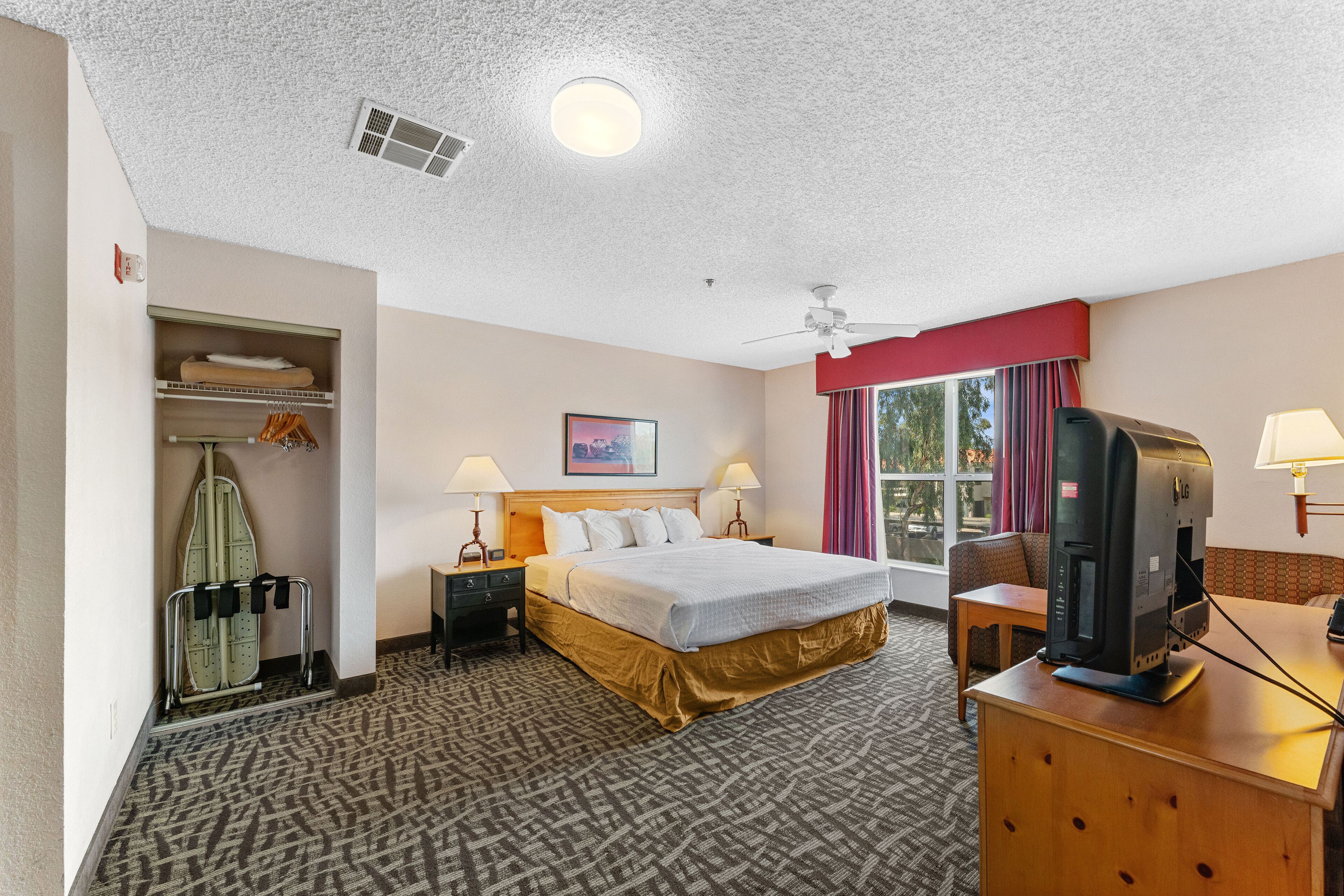 Una habitación de hotel con una cama, un escritorio con computadora, televisión, armario con ropa colgada, ventana con cortinas y ventilador de techo.
