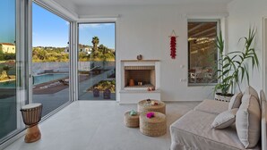 A modern living room with a large glass door leading to a pool, a fireplace, and a view of the surrounding landscape. - Villa Annassa Almyrida (CHANIA)