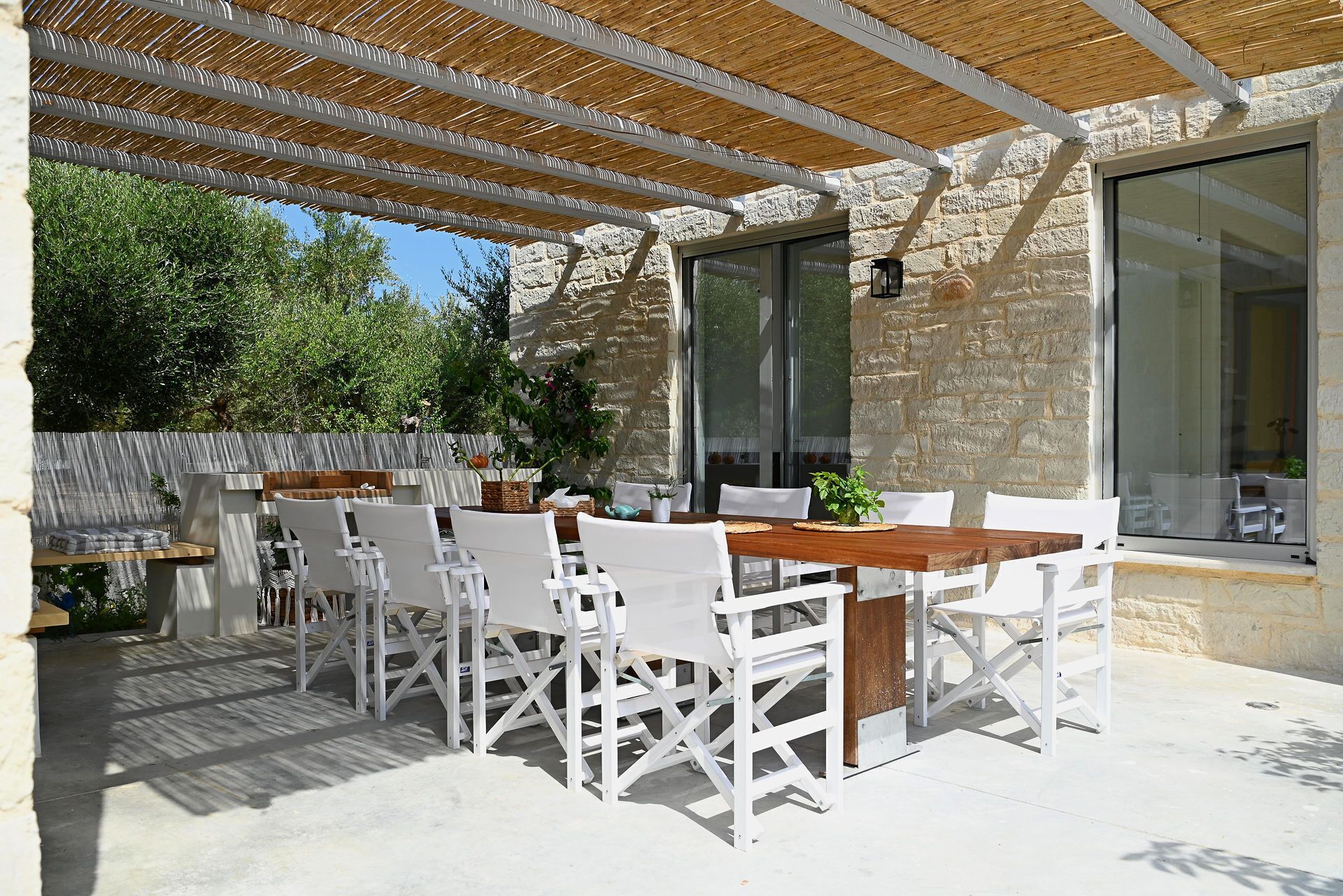 Eine Terrasse im Freien mit einem Holztisch und weißen Stühlen, einer Steinmauer und einer Glastür, die nach innen führt.