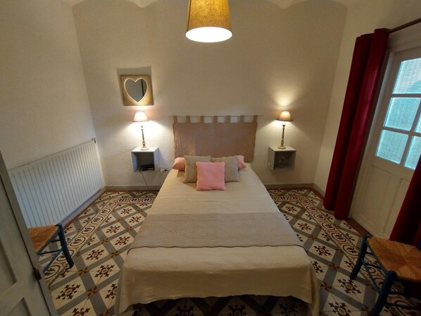 A hotel room with a bed, two bedside tables with lamps, a patterned floor, a radiator, and a window with curtains. - Village house, Gîte classé *** in the heart of the most beautiful sites in the Ardèche (Grospierres)