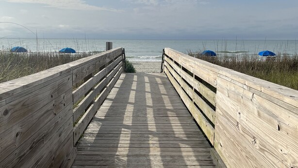Une promenade en bois menant à la plage, avec des parasols bleus et une vue sur l’océan.
