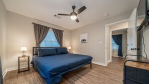 Una camera da letto con un letto, una ventola a soffitto, un comodino con una lampada, una finestra con tende e una porta che dà su un'altra stanza.