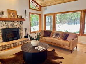 A cozy living room with a stone fireplace, a leather sofa, and large windows offering a view of a snowy landscape. - Lux, creekfront 8-acre retreat with hot tub (Nevada City)