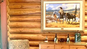 L’intérieur d’une cabane en rondins, avec un fauteuil à motifs, un meuble en bois orné d’objets décoratifs et un tableau représentant une calèche tirée par des chevaux.