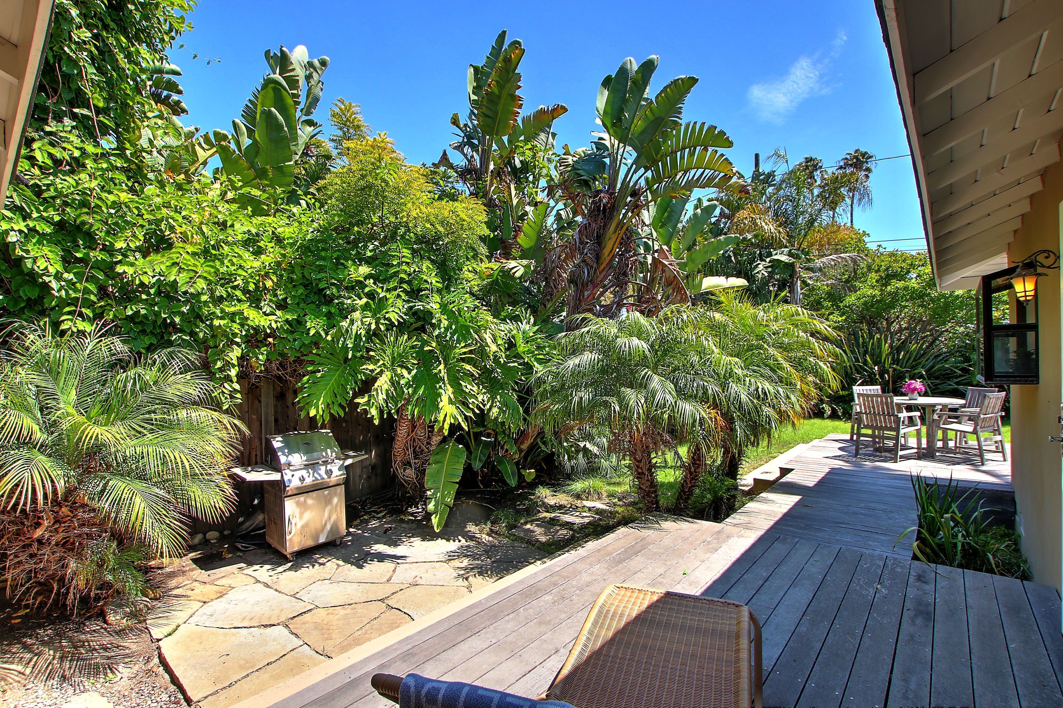 A wooden deck with a grill, surrounded by lush tropical plants and a patio table with chairs.