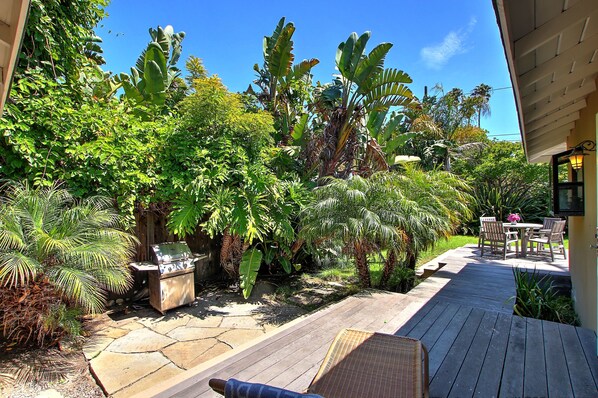 A wooden deck with a grill, surrounded by lush tropical plants and a patio table with chairs. - Little Palm Cottage - Steps from Shoreline Park, walk to beach, live like a local (Santa Barbara)