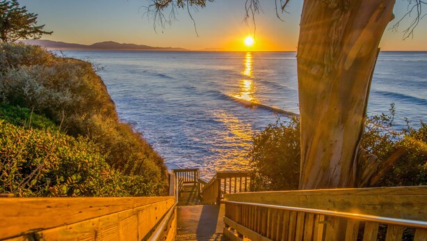 A wooden staircase leading down to a beach with a sunset in the background. - Little Palm Cottage - Steps from Shoreline Park, walk to beach, live like a local (Santa Barbara)
