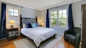 A bedroom with a bed, two bedside tables, a nightstand, a chair, and two windows with curtains. - Little Palm Cottage - Steps from Shoreline Park, walk to beach, live like a local (Santa Barbara)