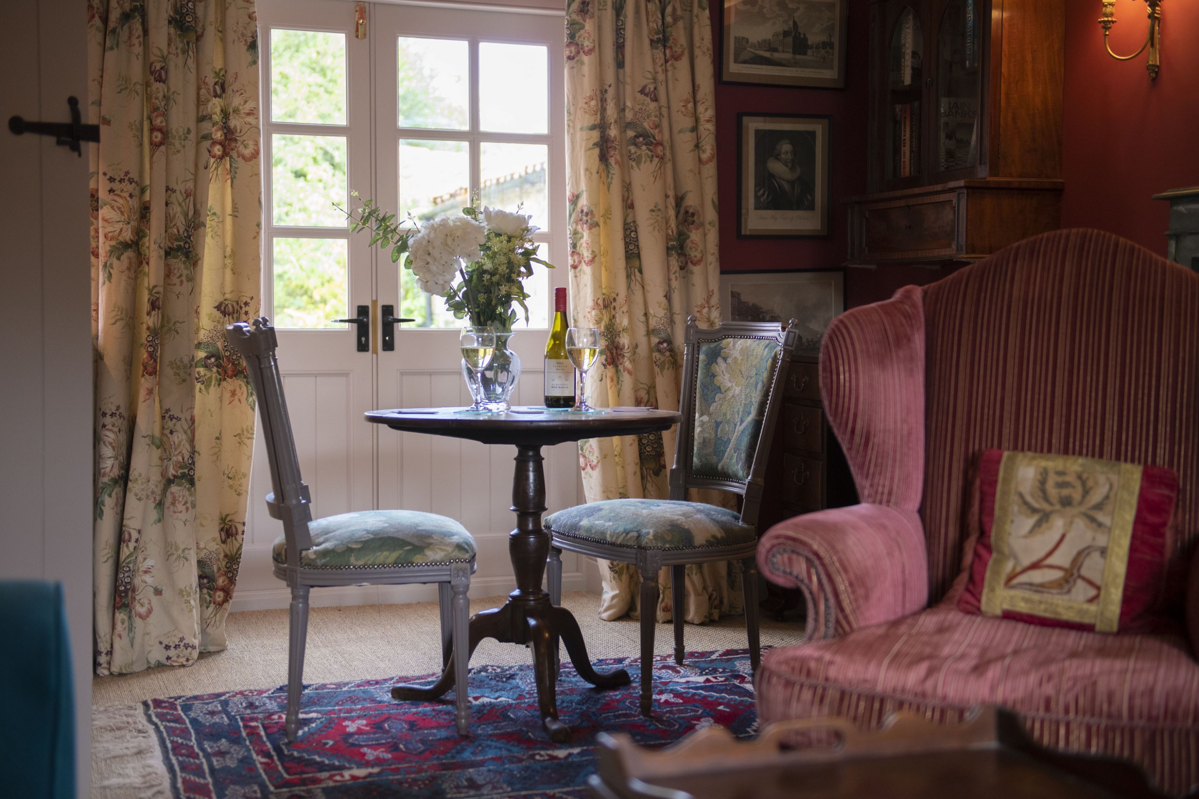 Une pièce comprenant une table à manger, deux chaises, un rideau à motifs floraux, une fenêtre avec vue et un fauteuil rouge recouvert de tissu.