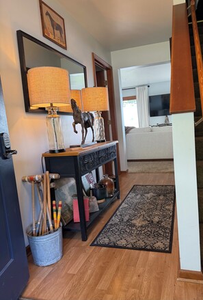 Una sala de estar con piso de madera, una consola decorada con objetos, un espejo grande y un televisor al fondo.