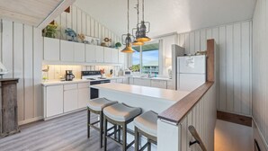 Une cuisine moderne avec des meubles blancs, un îlot central doté de tabourets de bar, un éclairage suspendu et une vue sur l’extérieur.