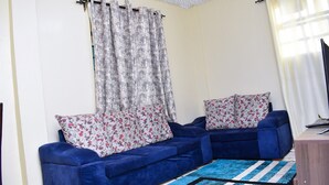 A living room with a blue sofa set, floral cushions, a patterned rug, and a television on a wooden stand. - KJ'S where Cosy Memories last. (Meru)