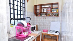 A compact kitchen with a sink, a microwave, a gas stove, and a wooden shelf with various kitchen items. - KJ'S where Cosy Memories last. (Meru)